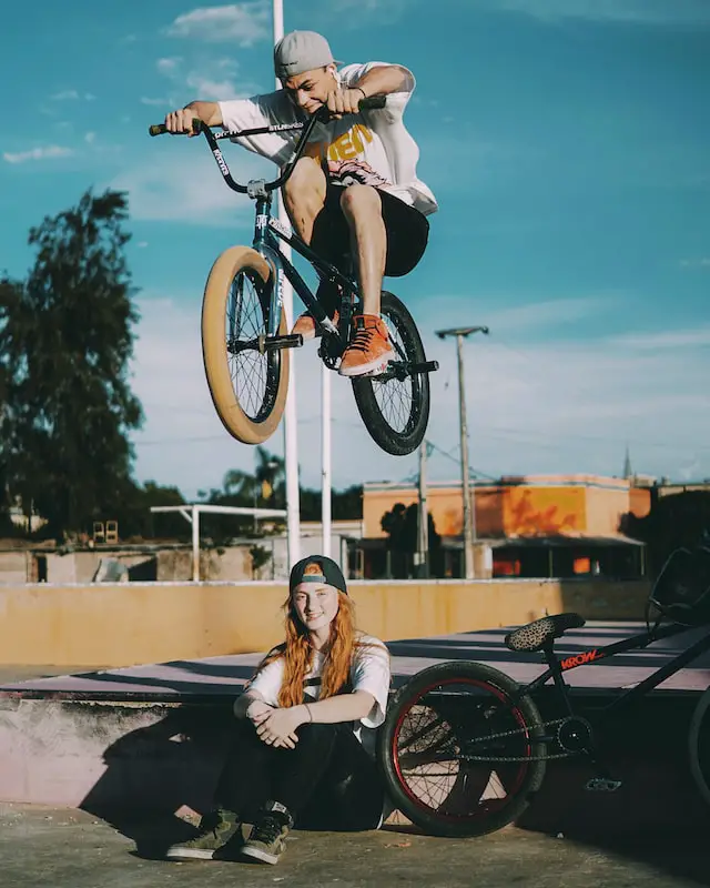 male bmx biker jumping over seated female bmxer